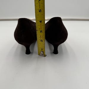 May include: A pair of dark brown suede pumps with a pointed toe and a small heel. The shoes are approximately 6 inches long, as measured by a yellow measuring tape.