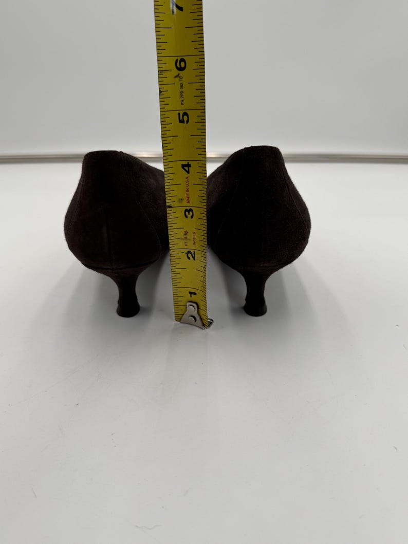 May include: A pair of dark brown suede pumps with a pointed toe and a small heel. The shoes are approximately 6 inches long, as measured by a yellow measuring tape.