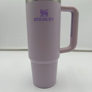 Op de afbeelding: Een lavendelkleurige Stanley-beker met een doorzichtig deksel en een bijpassende handgreep. Het Stanley-logo is paars op de voorkant gedrukt. De beker is ontworpen om dranken urenlang koud of warm te houden.