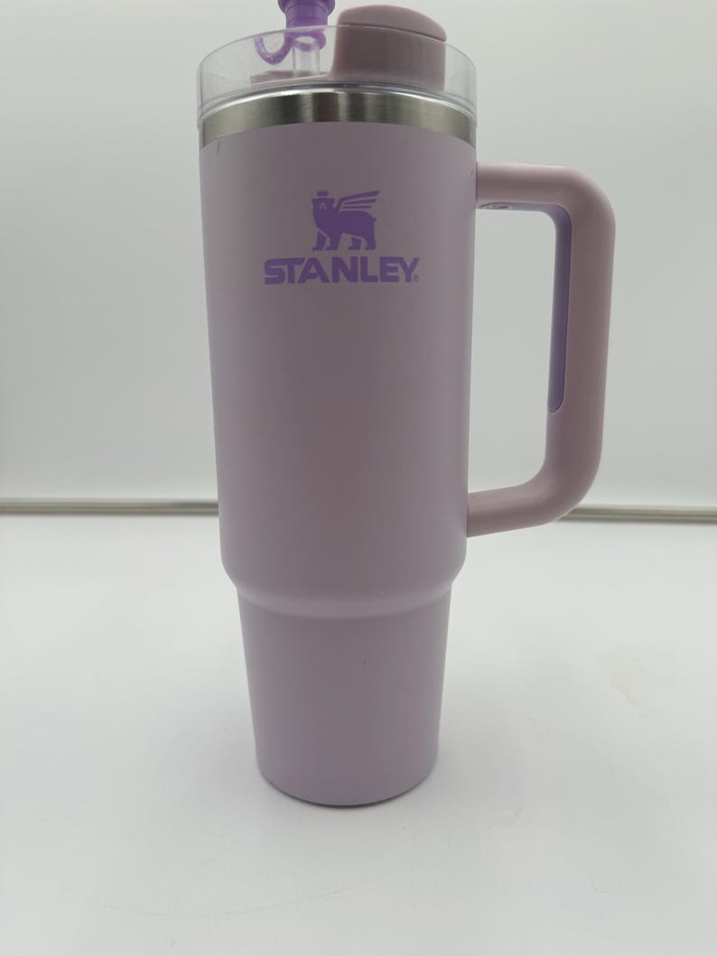 Op de afbeelding: Een lavendelkleurige Stanley-beker met een doorzichtig deksel en een bijpassende handgreep. Het Stanley-logo is paars op de voorkant gedrukt. De beker is ontworpen om dranken urenlang koud of warm te houden.