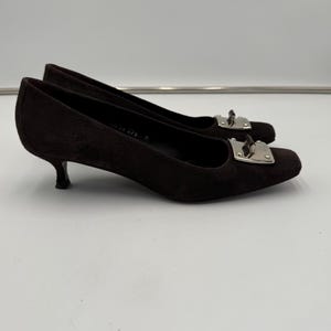 May include: A pair of dark brown suede heels with a square toe and a small, curved heel. Each shoe features a silver-colored metal buckle detail on the vamp. The shoes are set against a plain white background.
