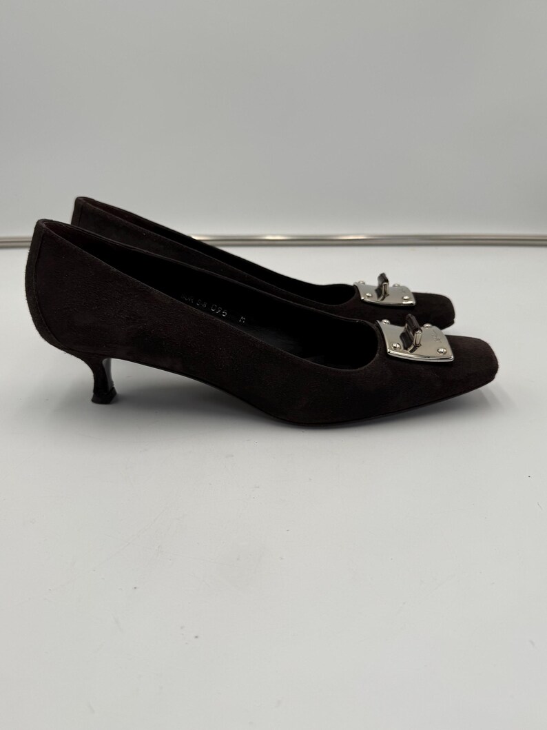 May include: A pair of dark brown suede heels with a square toe and a small, curved heel. Each shoe features a silver-colored metal buckle detail on the vamp. The shoes are set against a plain white background.