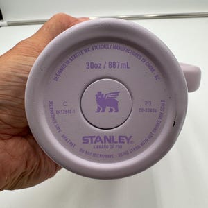 Op de afbeelding: De bodem van een lichtpaarse Stanley-beker. De bodem van de beker heeft de tekst "30oz / 887mL". De tekst "Designed in Seattle WA, Ethically Manufactured in China - BC" is ook zichtbaar.
