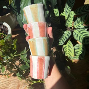 Può includere: Una pila di quattro tazze cilindriche in ceramica con strisce verticali. Due tazze presentano strisce bianche e color cuoio con sottili accenti verdi, mentre le altre due tazze hanno strisce bianche e rosso-arancio con finitura lucida.