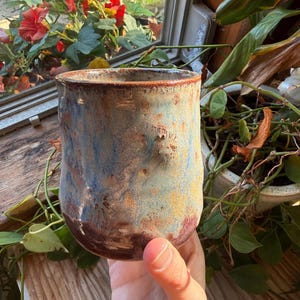 Op de afbeelding: Een handgemaakte keramische mok met een unieke, getextureerde glazuur. De mok heeft een mix van blauwe, bruine en rood-paarse tinten, wat een rustieke en artistieke uitstraling creëert. De cilindrische vorm is ideaal voor warme dranken.