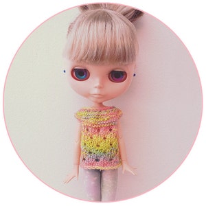 May include: A doll with blonde hair and blue eyes wearing a rainbow crochet top and patterned leggings.
