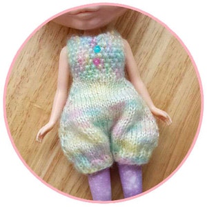 May include: A doll wearing a pastel-colored knitted jumpsuit with a beaded design on the chest. The jumpsuit has a loose fit and is made of a soft, textured yarn.