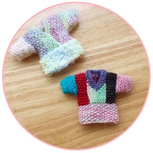 Może przedstawiać: Dwa miniaturowe swetry dziane, jeden w paski tęczowe, a drugi w paski wielokolorowe, na drewnianej powierzchni.