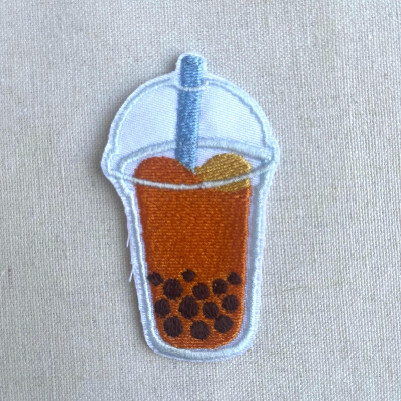 May include: An embroidered patch of a boba tea cup with a straw, brown tapioca pearls, and a white lid. The boba tea is orange and yellow.