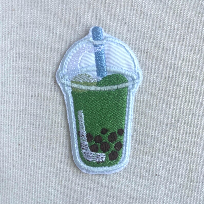 May include: An embroidered patch of a green bubble tea with a white straw and a white lid. The bubble tea has brown tapioca pearls.