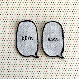 May include: Two white embroidered patches with black outlines in the shape of speech bubbles. The left patch says "ばか" in Japanese. The right patch says "BAKA" in English.
