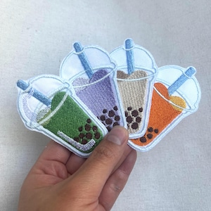 May include: Four embroidered iron-on patches of bubble tea in different colors: green, purple, brown, and orange. Each patch features a white cup with a straw and boba pearls.