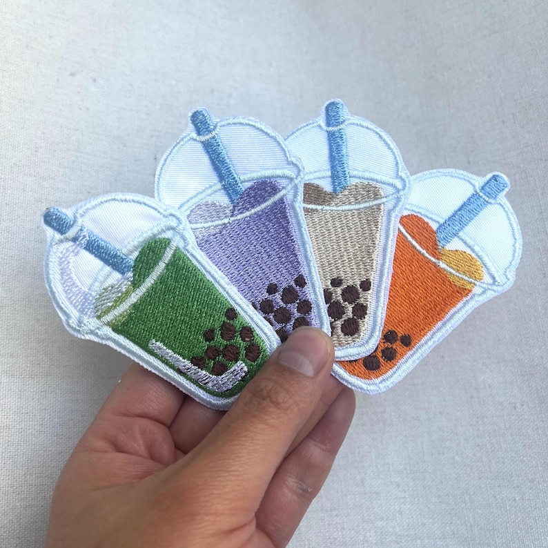 May include: Four embroidered iron-on patches of bubble tea in different colors: green, purple, brown, and orange. Each patch features a white cup with a straw and boba pearls.