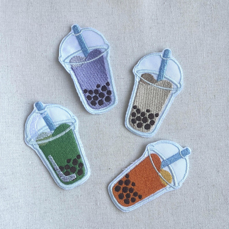 May include: Four embroidered iron-on patches of bubble tea drinks with different colored liquids and boba pearls. The patches are on a light gray fabric background.