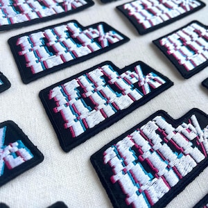 May include: A set of black embroidered iron-on patches with a white, pink, and blue glitch effect design. The patches feature the text "111%" in a pixelated font.