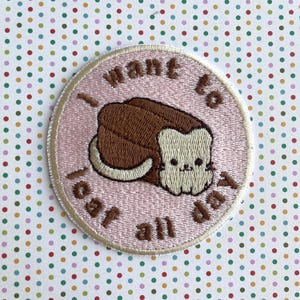 May include: A pink embroidered patch with the text "I want to eat all day" and a cartoon illustration of a brown loaf of bread with a white cat face.