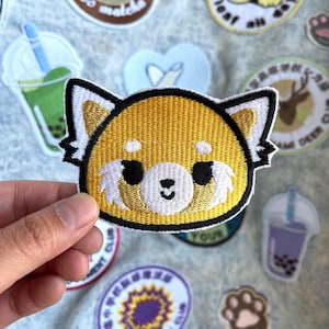 May include: Embroidered patch of a red panda with a yellow and white face, black eyes, and a white muzzle. The patch has a black border and is surrounded by other patches with various designs, including a bubble tea and a deer.