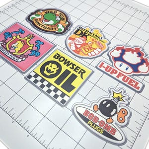 Könnte beinhalten: Eine Sammlung farbenfroher, rechteckiger Aufkleber mit Cartoon-Charakteren und Text. Designs umfassen "Wild Yoshi Sanctuary", "Princess Peach", "Burning DK", "Bowser Oil", "I-Up Fuel" und "Bob-Omb Plugs."