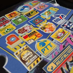 Peut inclure: Une collection d'autocollants rectangulaires et ovales colorés avec divers personnages de dessins animés et logos. Les autocollants incluent du texte tel que "Mushroom Engines", "Yoshi's Egg", "Burning DK" et "Bullet Bill Speed Trial".