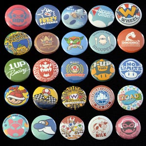Könnte beinhalten: Eine Sammlung von fünfundzwanzig runden Ansteckbuttons mit verschiedenen bunten Grafikdesigns, darunter Zeichentrickfiguren, Textlogos und Embleme. Die Buttons sind in einem fünf-mal-fünf Raster auf einem schwarzen Hintergrund angeordnet.