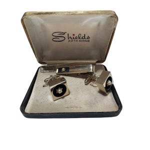 May include: A vintage gold-tone cufflink and tie clip set with cultured pearls, presented in a black and gold box. The cufflinks have a square design with a black centre and a pearl. The tie clip has a black centre and a pearl. The box is labelled "Shields Fifth Avenue."