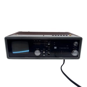 May include: Vintage portable TV/radio with a digital clock. The device has a dark wood-grain top and a black front panel with silver knobs. It features a small screen on the left and a radio dial on the right. A black power cord extends from the bottom.