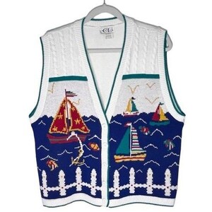 May include: A white and blue knit button-down vest featuring a nautical pattern with sailboats, fish, and a white picket fence design along the bottom hem. The garment has a V-neckline and cable knit detailing on the upper white section.