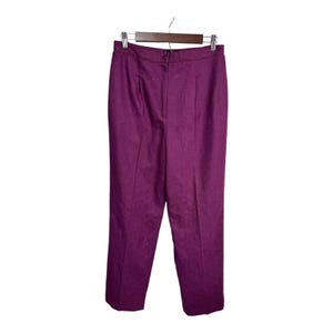 May include: A pair of solid purple trousers hanging on a wooden hanger. The trousers are made of a textured fabric and feature a button closure and pleats at the waist. The trousers are a vibrant shade of purple.