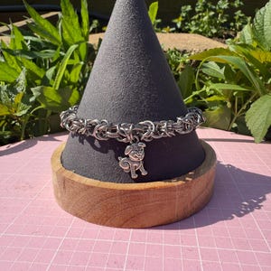 May include: A silver-toned chainmail bracelet with a dog charm is displayed on a gray cone-shaped jewelry stand. The stand sits on a wooden base, set against a pink grid background with greenery in the background.