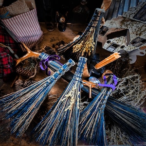 May include: Three decorative brooms with blue and natural bristles, tied with purple ribbon. The brooms are decorated with dried wheat and other natural elements. The brooms are arranged on a wooden surface with other rustic decor.
