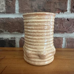 May include: A handmade, cylindrical ceramic vase with a textured, ridged design. The vase is a light beige color with brown streaks, and it sits on a wooden surface. The background features a brick wall.