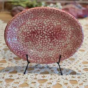 May include: An oval-shaped ceramic serving dish with a speckled red and white pattern. The dish is displayed on a black metal stand, showcasing its design. The background features a brick wall and decorative elements, highlighting the dish's aesthetic appeal.