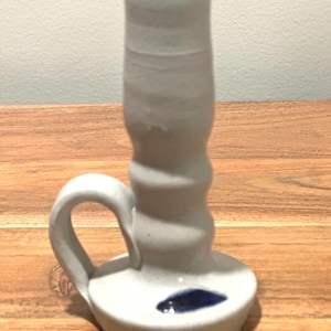 May include: A hand-thrown ceramic candle holder with a light gray speckled finish. The piece features a cylindrical stem with a rounded handle and a circular base. A dark blue glaze accents the top rim of the stem and a small spot on the base.