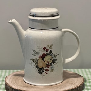May include: Cream-colored ceramic coffee pot with a lid and handle. The pot features a fruit and berry design in shades of red, green, and yellow. The pot is sitting on a wooden slice.