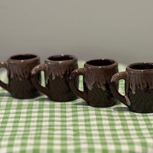 May include: Four small ceramic mugs arranged in a row. Each mug features a dark brown base with a textured, basket-weave pattern and a lighter tan glaze dripping from the rim down the sides. Each mug has a single handle on the side.