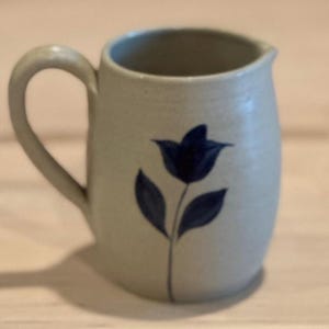 May include: A small, light-colored ceramic pitcher with a handle. The pitcher is decorated with a dark blue floral design. The pitcher is likely used for serving liquids.