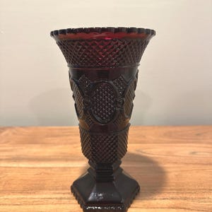 May include: A ruby red glass vase with a diamond pattern and ornate oval designs. The vase has a flared top with a scalloped edge and a hexagonal base. The vase is approximately 9 inches tall.