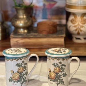 May include: Two white ceramic mugs with handles, featuring a yellow rose illustration and the word "Rosa". Each mug has a teal rim and lid with a botanical design. The mugs are part of a tea set.