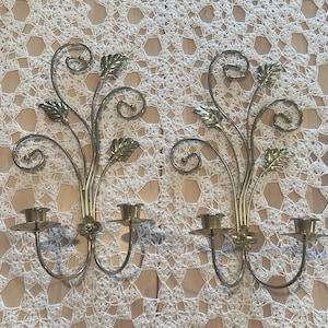 May include: A pair of vintage brass wall sconces. Each sconce features a floral design with leaves and curling stems, and two candle holders. The sconces are displayed against a white lace background.