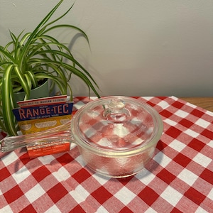 May include: A clear glass Range-Tec stovetop cooking pot with a lid, sitting on a red and white checkered tablecloth. The pot has a handle and is next to a package with the text "The Key to Modern Cooking". A green plant is in the background.