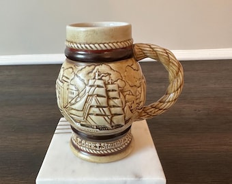 May include: A beige ceramic mug with a nautical theme. The mug features a detailed relief of a sailing ship and buildings. The handle is designed to look like twisted rope. The base of the mug reads "Bark Staysail".