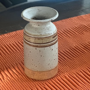 May include: A cylindrical ceramic vase with a speckled off-white glaze, featuring a flared rim and horizontal brown bands around the neck. The base of the vase is unglazed, showing a textured, earthy brown clay finish.