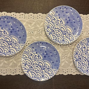 May include: A set of four round ceramic plates featuring a blue and white hand-painted pattern of stylized waves and scattered dots. The design covers the lower portion of each plate with concentric arcs, while the upper portion displays blue dots.