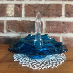 May include: A vintage, deep teal glass basket with a clear, arched handle. The basket has a ruffled edge and sits on a white, crocheted doily. The background features a brick wall and a wooden surface.