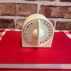 May include: Cream-colored Pyrex kitchen timer with a red dial and black numbers. The timer has a white knob and the words "KITCHEN TIMER" are printed on the dial. The timer is sitting on a red surface.