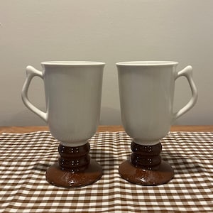 May include: Two white ceramic mugs with a unique design. The mugs have a cylindrical shape with a curved handle. They sit on a brown, pedestal-style base with a stacked, rounded design. The mugs are placed on a brown and white checkered tablecloth.