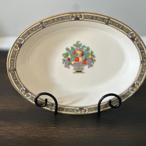 May include: An oval ceramic serving dish with a cream-colored center featuring a central fruit basket illustration. The rim is decorated with a dark blue and gold patterned border. The dish is supported by a black metal wire display stand.