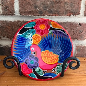 May include: A hand-painted, decorative ceramic bowl featuring a vibrant bird design with blue, pink, orange, and green colors. The bowl is supported by a black metal stand, showcasing a folk art style. The rim is red.
