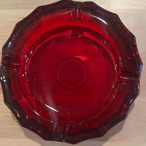 May include: A circular, deep red glass ashtray featuring a faceted, scalloped edge design. The center of the ashtray displays a circular embossed emblem with a decorative beaded border, providing a textured contrast to the smooth, translucent red glass surface.