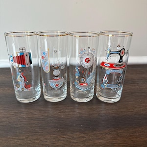 May include: Set of four vintage highball glasses with gold rims. Each glass features a different colorful design: an accordion, a decorative vase, a clock, and a sewing machine. The glasses are clear with a gold rim.
